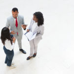Diverse business team meeting in office hallway. Business man and women standing in circle, holding tablet and documents and talking. Teamwork concept