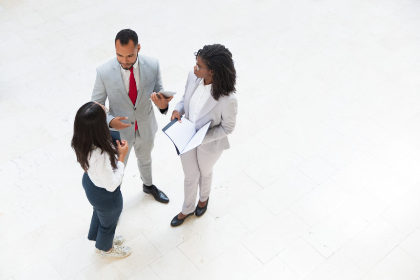 Diverse business team meeting in office hallway Diverse business team meeting in office hallway. Business man and women standing in circle, holding tablet and documents and talking. Teamwork concept